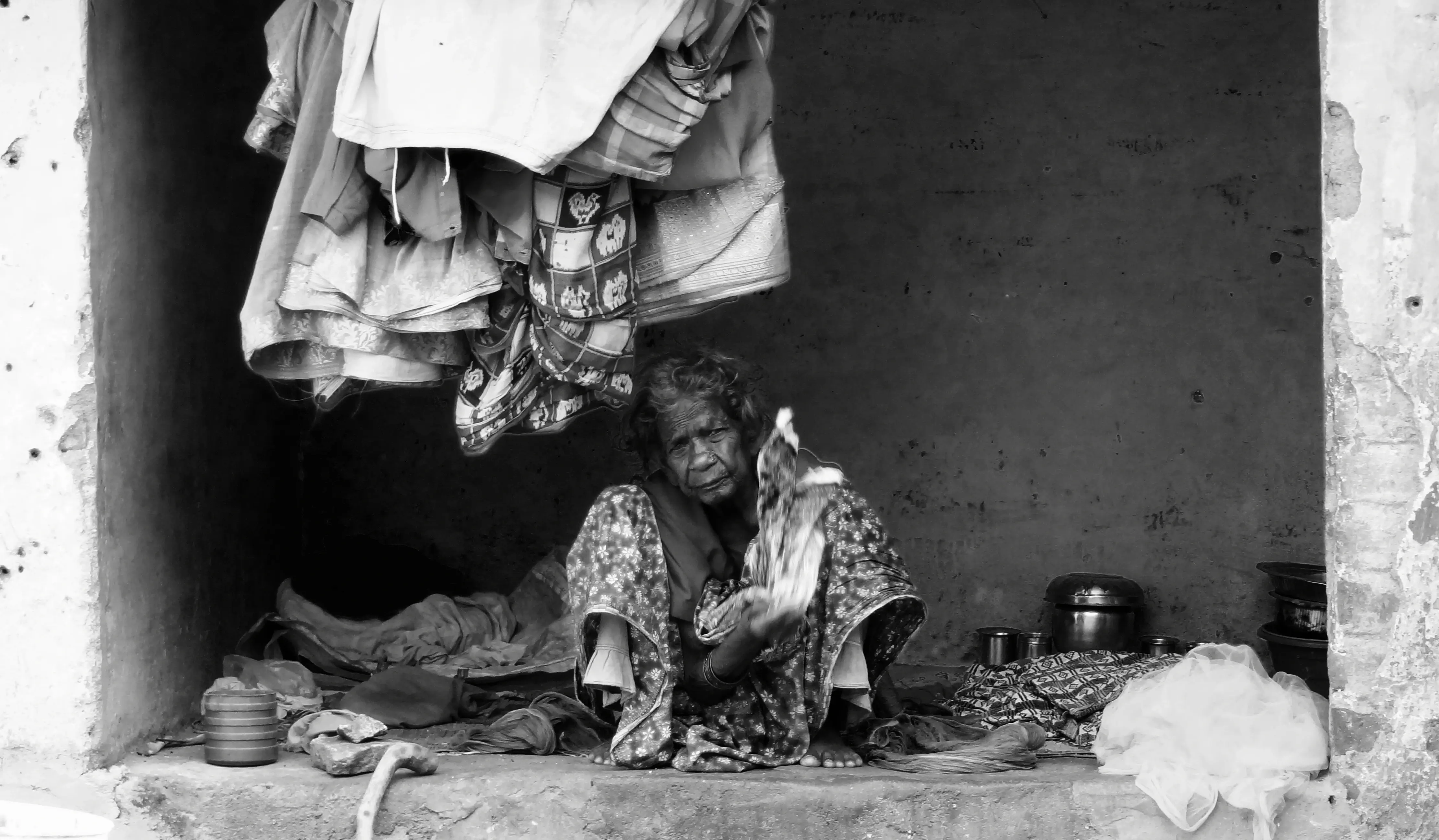 Elderly woman sitting under hanging clothes in a small shelter.