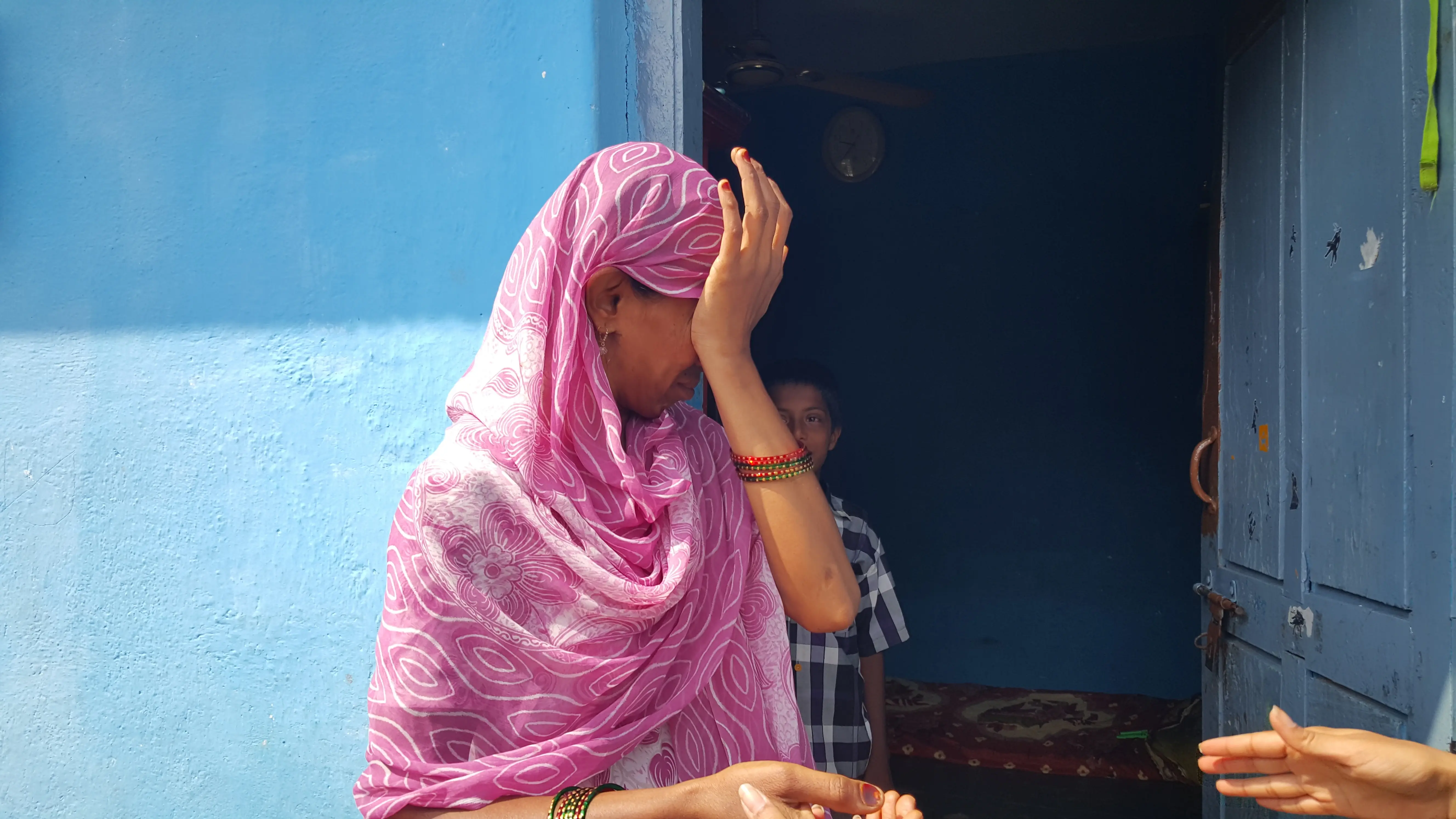 Woman in a pink headscarf wiping her face outside a blue doorway.